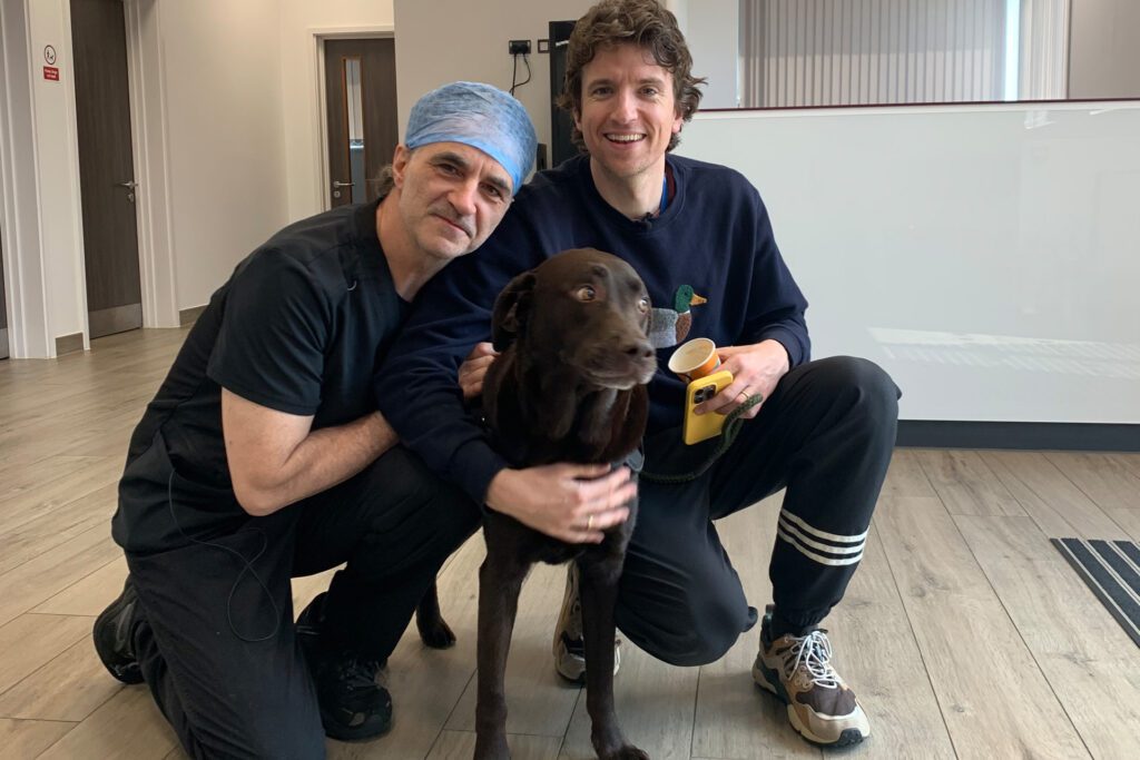 Professor Noel Fitzpatrick crouching next to a chocolate Labrador and their owner in reception