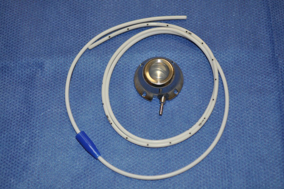 A Vascular Access Port prior to insertion beneath the skin. The metal disk has a central resin chamber through which the drugs are injected. One end of the white tubing is connected to this chamber, the other end is inserted, and left, in a blood vessel. This device removes the need for constant restraint and leg injections.